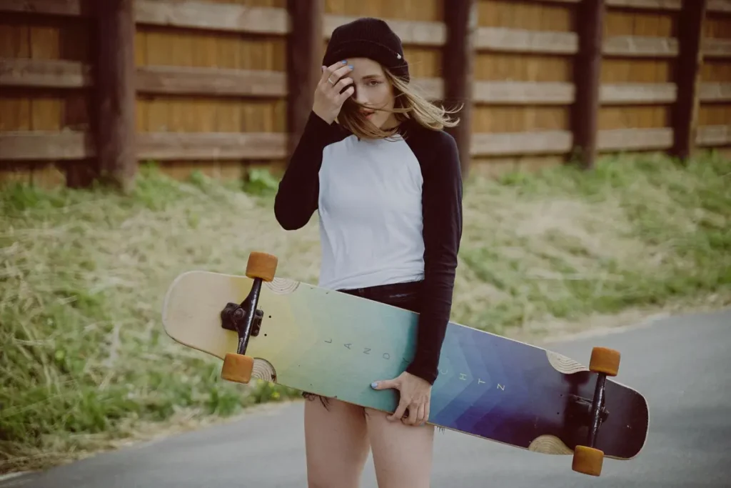 A young woman holding a colourful longboard by a wooden fence