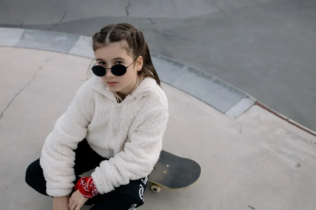 A teenage girl in sunglasses sitting on a skateboard at a skatepark