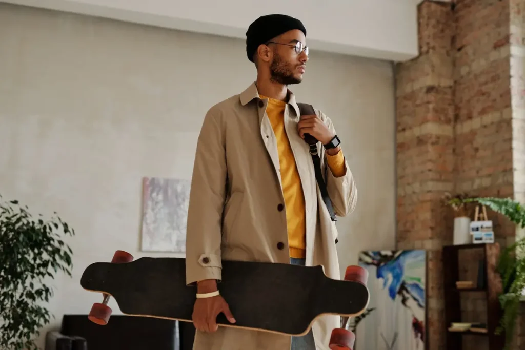 A man in a trench coat and hat holding a skateboard indoors