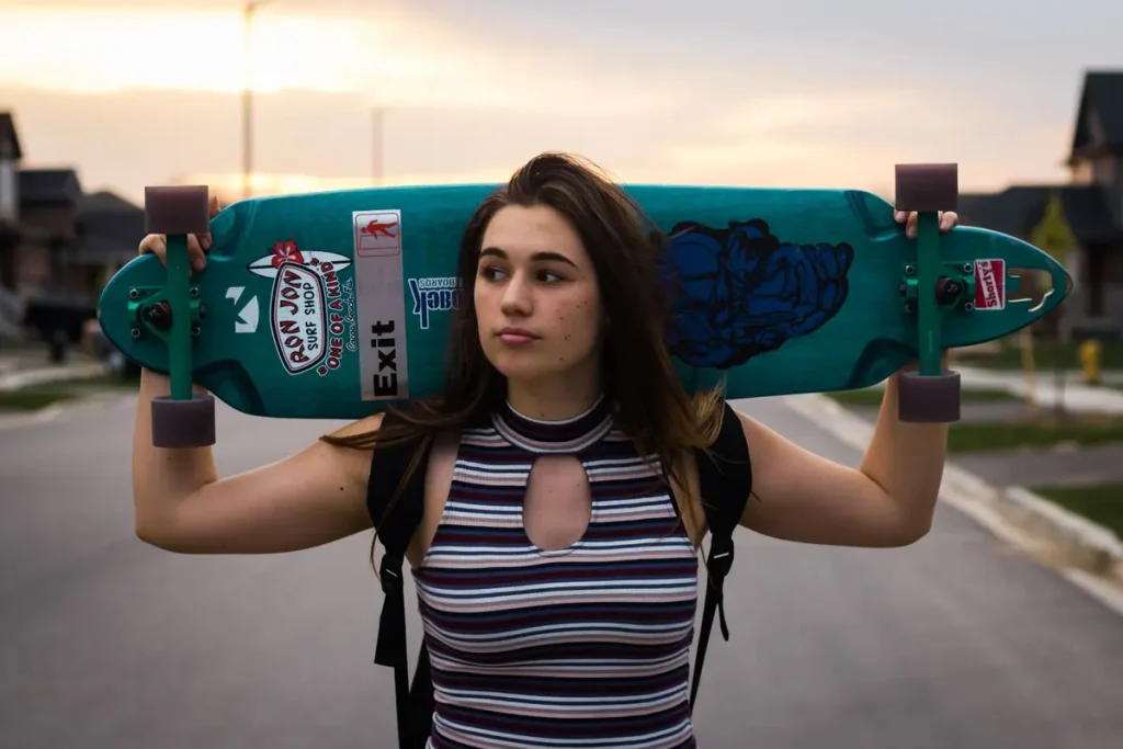 A teenage girl carrying a longboard on her shoulders