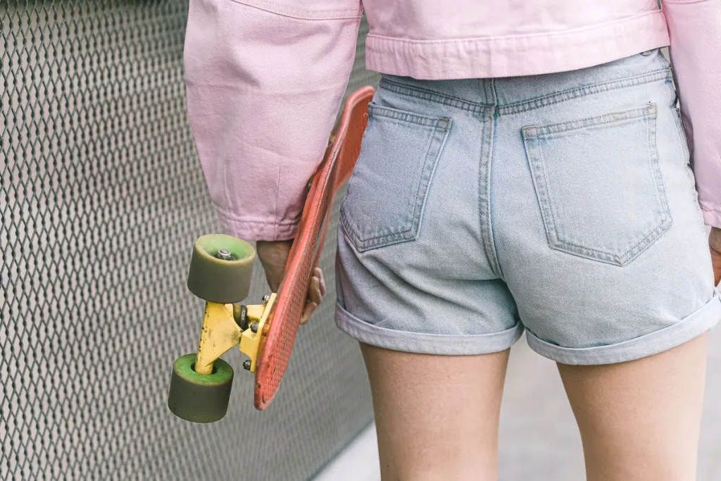 A person in denim shorts and a pink jacket holding a skateboard