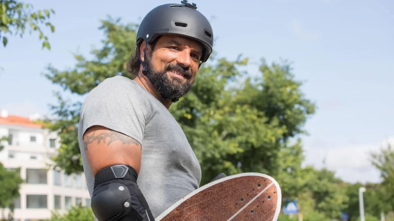 A smiling man in a helmet and elbow pads holding a skateboard
