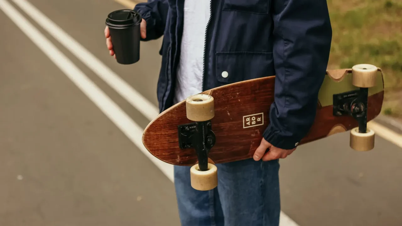 A young man holding a longboard and a coffee on a street